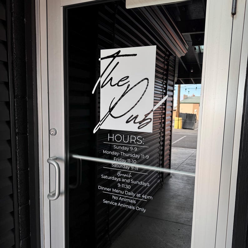 May include: A glass door with a white sign displaying the name "The Dub" in cursive lettering. The sign also lists opening hours, including brunch times on Saturdays and Sundays. The door has a silver handle and is set in a black frame.