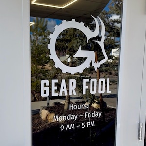 May include: A glass door featuring a white logo and text. The logo is a gear combined with an animal head. The text reads "GEAR FOOL" and "Hours: Monday - Friday 9 AM - 5 PM". The background shows trees and a blue sky.
