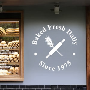 Puede incluir: Texto blanco en una pared gris que dice "Baked Fresh Daily Since 1975". Un rodillo blanco y una espiga de trigo se cruzan en el centro del c&iacute;rculo.