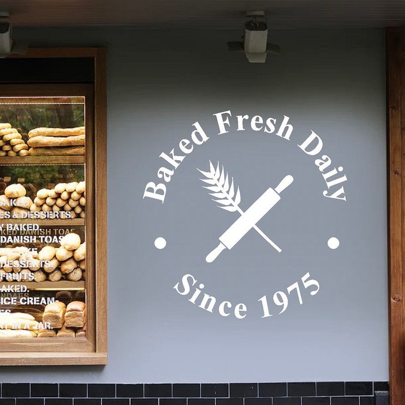 Puede incluir: Texto blanco en una pared gris que dice "Baked Fresh Daily Since 1975". Un rodillo blanco y una espiga de trigo se cruzan en el centro del c&iacute;rculo.