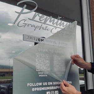 Op de afbeelding: Een etalage met de tekst "Premium Graphic Co" in een gestileerd lettertype. De woorden "Custom Vinyl" en "Scan to" zijn ook zichtbaar. Een QR-code en de woorden "Shop Our Website" en "Follow Us On Social Media @Premiumgraph" zijn ook zichtbaar.
