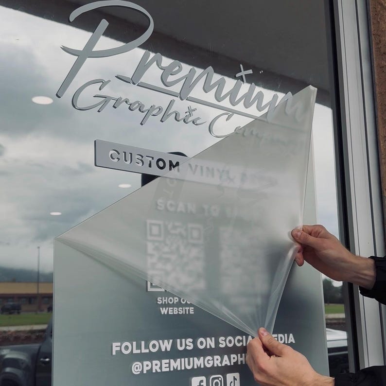 May include: A shop window displaying the text "Premium Graphic Co" in a decorative font. The words "Custom Vinyl" and "Scan to" are also visible. A QR code and the words "Shop Our Website" and "Follow Us On Social Media @Premiumgraph" are also visible.
