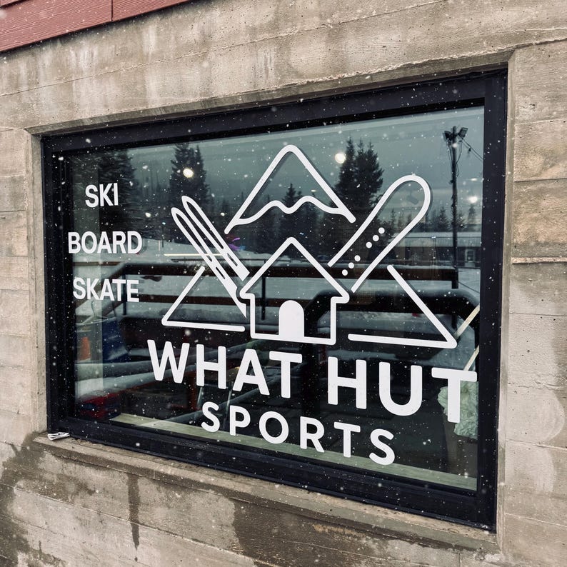 May include: A shop window with a black frame features a white graphic logo for What Hut Sports. The logo includes a mountain, a house, skis, and a snowboard. The words "SKI", "BOARD", "SKATE", and "SPORTS" are also visible.