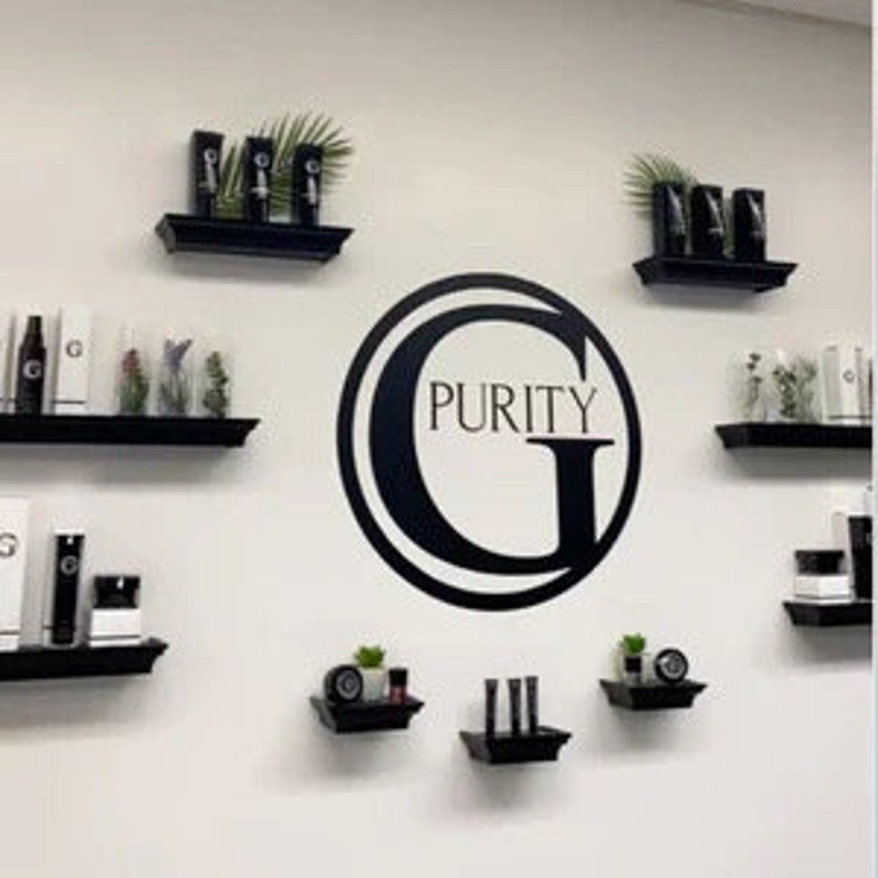 Puede incluir: Una pared blanca con estantes negros que muestran varios productos de belleza. Un c&iacute;rculo negro grande con el texto "PURITY" y una "G" estilizada en su interior est&aacute; en la pared.