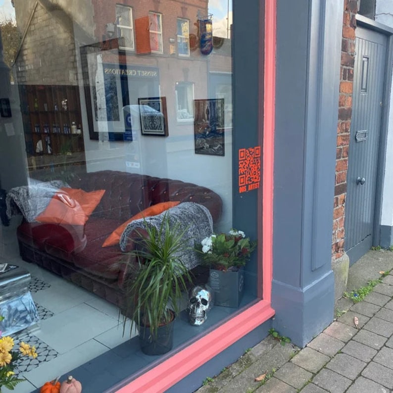 Peut inclure: Une vitrine de magasin pr&eacute;sente un canap&eacute; en cuir bordeaux avec des coussins oranges et une couverture grise. Un pot de plante, un cr&acirc;ne et des fleurs sont sur le rebord. La fen&ecirc;tre refl&egrave;te des &oelig;uvres encadr&eacute;es et le texte "SUNSET CREATIONS".