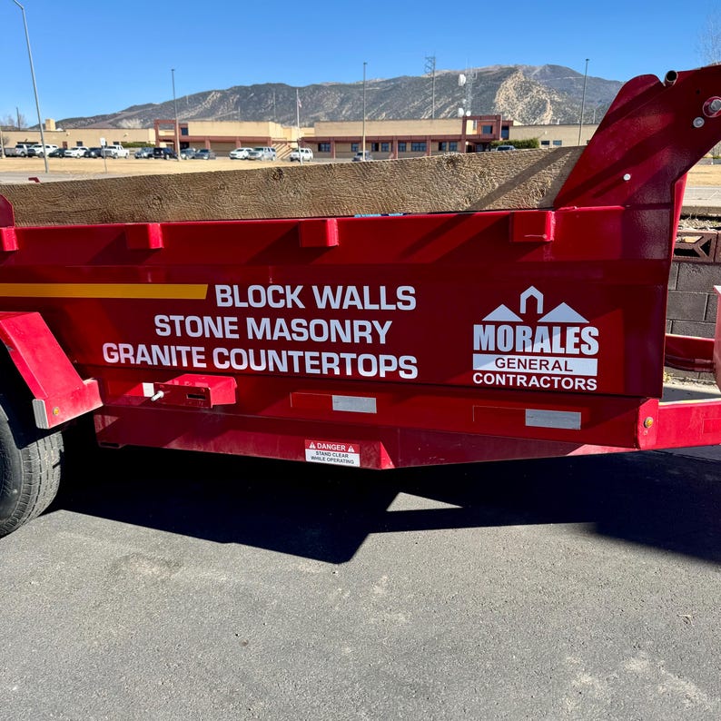 May include: A red construction trailer with white text reading "BLOCK WALLS, STONE MASONRY, GRANITE COUNTERTOPS." The Morales General Contractors logo is on the right. The trailer is filled with brown material.