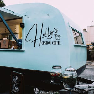 Può includere: Un rimorchio vintage azzurro con la scritta "Holly's Custom Coffee" in corsivo nero. Due distributori di bevande in vetro sono posizionati sotto la finestra del rimorchio. Un piccolo cartello lavagna recita "Pop Fizz".
