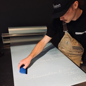 Peut inclure: Une personne utilise une raclette bleue pour appliquer un graphisme sur une surface bleu clair. Le graphisme comprend le texte "YOU LOOK INCREDIBLE. NOW GO GRAB A DRINK!" et les noms "Alexandria & Alan Tuthill" avec la date "10.07.23".