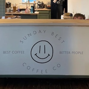 Puede incluir: Un letrero blanco con texto negro que dice "Sunday Best Coffee Co." con un logotipo de cara sonriente y el texto "Best Coffee" y "Better People".