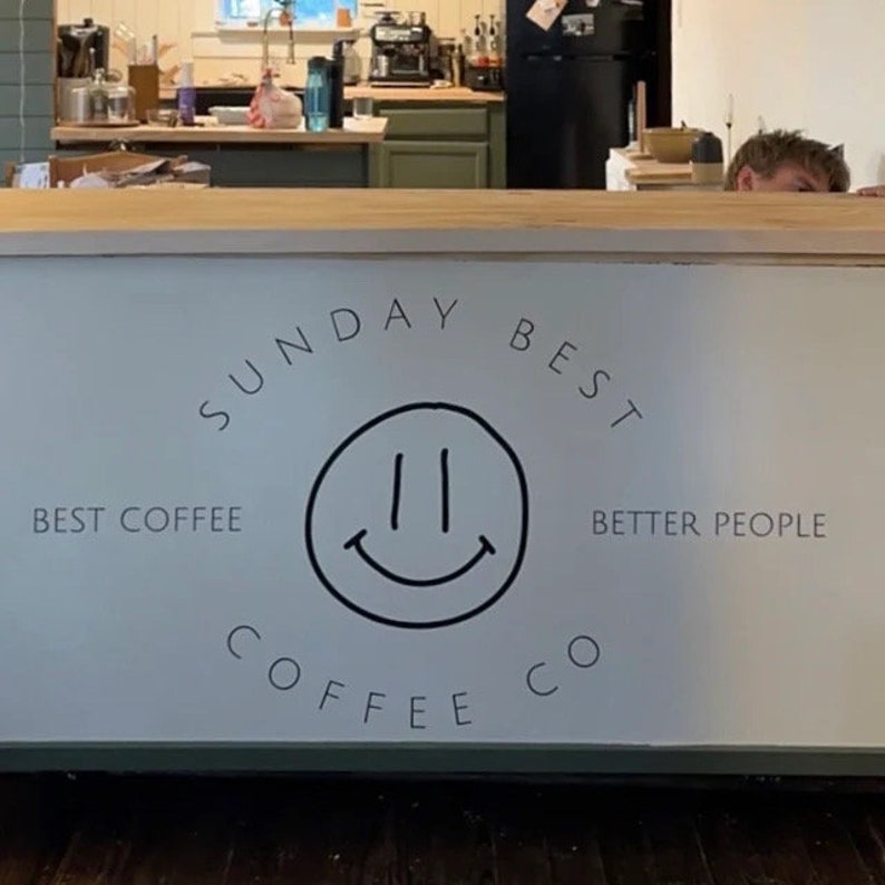 Puede incluir: Un letrero blanco con texto negro que dice "Sunday Best Coffee Co." con un logotipo de cara sonriente y el texto "Best Coffee" y "Better People".