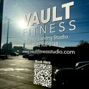Peut inclure: Une porte vitr&eacute;e avec le texte blanc "VAULT FITNESS" et "Private Training Studio". Un num&eacute;ro de t&eacute;l&eacute;phone et une adresse de site Web sont en dessous. Un code QR et les mots "Book Here" sont dans le coin inf&eacute;rieur droit. L'arri&egrave;re-plan montre une sc&egrave;ne de rue.