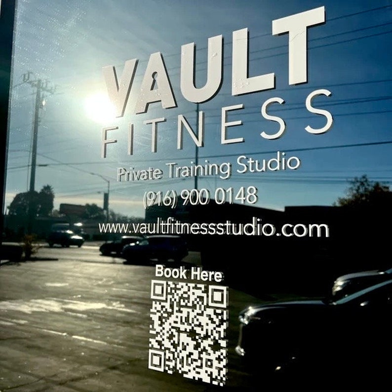 Peut inclure: Une porte vitr&eacute;e avec le texte blanc "VAULT FITNESS" et "Private Training Studio". Un num&eacute;ro de t&eacute;l&eacute;phone et une adresse de site Web sont en dessous. Un code QR et les mots "Book Here" sont dans le coin inf&eacute;rieur droit. L'arri&egrave;re-plan montre une sc&egrave;ne de rue.