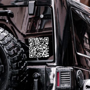 Puede incluir: Un coche negro con una pegatina de código QR blanco en la ventana trasera. El coche tiene una rueda de repuesto en la parte trasera.