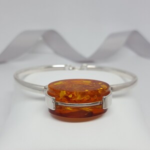 May include: A silver bangle bracelet featuring a large oval amber stone set in the centre. The amber is a warm, honey colour with inclusions.