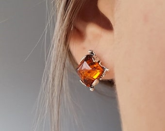 May include: A silver-toned stud earring with a square, amber-colored gemstone. The gemstone is faceted, catching the light and displaying a warm, golden hue. The earring has a decorative, textured setting. Jewelry.