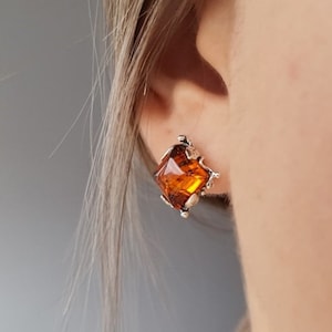 May include: A silver stud earring featuring a square, amber gemstone. The gemstone is faceted, reflecting light and displaying a warm, golden hue. The earring has a decorative, textured setting. Jewellery.