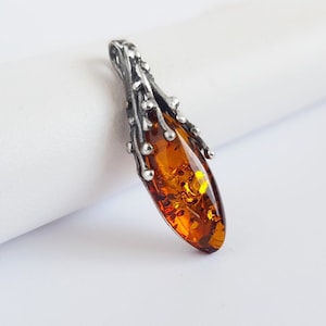 May include: A silver pendant featuring an amber-coloured, teardrop-shaped stone. The silver setting has a textured, organic design with small, round accents. The amber stone is translucent, with visible inclusions, set against a white background.