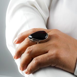 May include: A statement ring featuring a large, dark, polished stone set in a silver band. The ring is worn on a hand, with a white shirt in the background. The stone has a deep, black hue and a smooth surface.