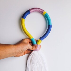 Peut inclure: Une couronne de laine colorée avec un pompon blanc suspendu en bas. La couronne est faite de laine bleue, jaune, rose et turquoise.