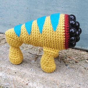 Puede incluir: Una criatura amarilla de crochet con rayas azules y rojas y ojos de cuentas negras. La criatura tiene cuatro patas y est&aacute; de pie sobre una superficie marr&oacute;n.