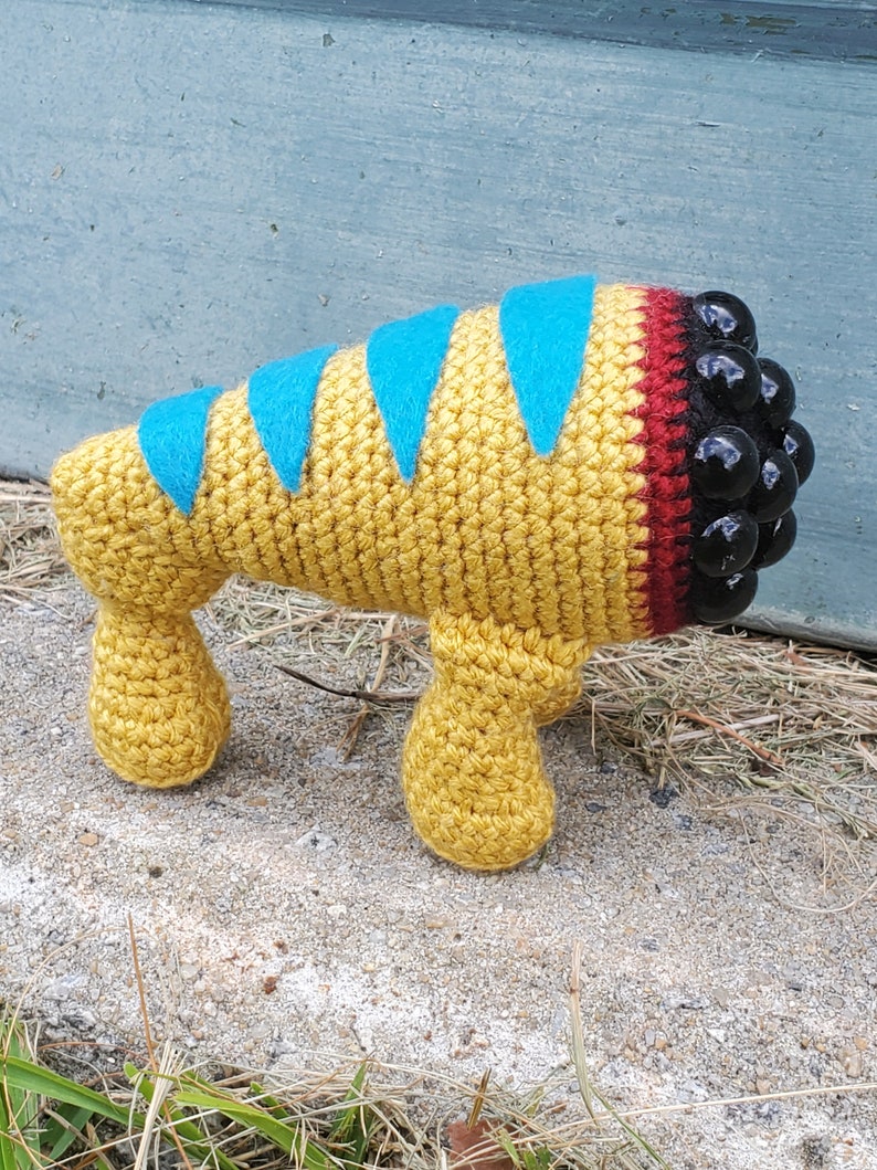 Puede incluir: Una criatura amarilla de crochet con rayas azules y rojas y ojos de cuentas negras. La criatura tiene cuatro patas y est&aacute; de pie sobre una superficie marr&oacute;n.