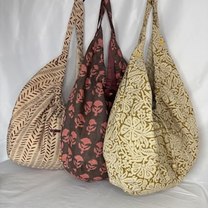 May include: Three fabric shoulder bags with different patterns. One bag is beige with a maroon chevron design, another is brown with pink floral prints, and the third is beige with a green floral pattern. Each bag has a long strap.
