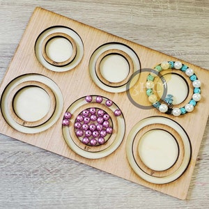 May include: Wooden board with five circular cutouts for measuring jewelry. The cutouts are labeled 6.5 inches, 7 inches, 8 inches, 8.5 inches, and 9 inches. A turquoise and white beaded bracelet is in the 8-inch cutout. A pile of purple beads is in the 8.5-inch cutout.