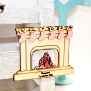 Puede incluir: Adorno navideño de madera con forma de chimenea con calcetines rosas con las etiquetas "DAD", "MOM", "TY", "ZAI", "NY" e "IZZY". La chimenea tiene un fuego recortado y el texto "Thompson's Christmas 2020".
