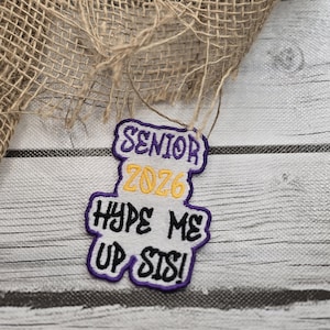 May include: A white embroidered patch with a purple border and the words "SENIOR 2026 HYPE ME UP SIS!". The text is in purple, yellow, and black. The patch is attached to a piece of twine and set against a burlap and wood background.