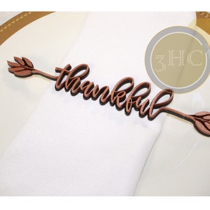 May include: A wooden napkin ring with the word "thankful" in a cursive font. The word is attached to arrow-shaped ends with leaf details. The napkin ring is brown and rests on a white napkin and a gold-rimmed plate.