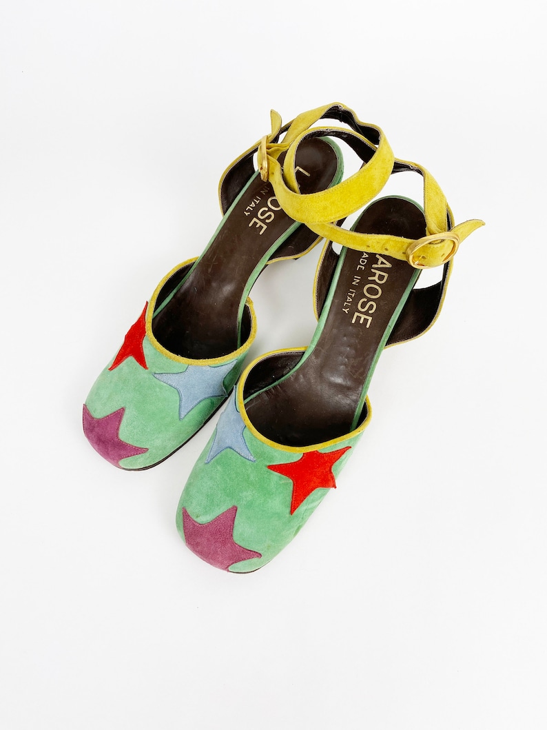 May include: A pair of green suede heeled shoes with a yellow ankle strap and a star pattern. The shoes are made in Italy by the brand "La Rose".