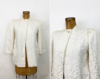 May include: A white, embroidered jacket with a mandarin collar and long sleeves. The jacket has a single button closure and two pockets.