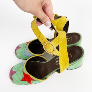 May include: Green suede heeled sandals with a yellow ankle strap and a star design. The sandals are made in Italy and have a brown leather insole. The brand name "L'AISE" is printed on the insole.