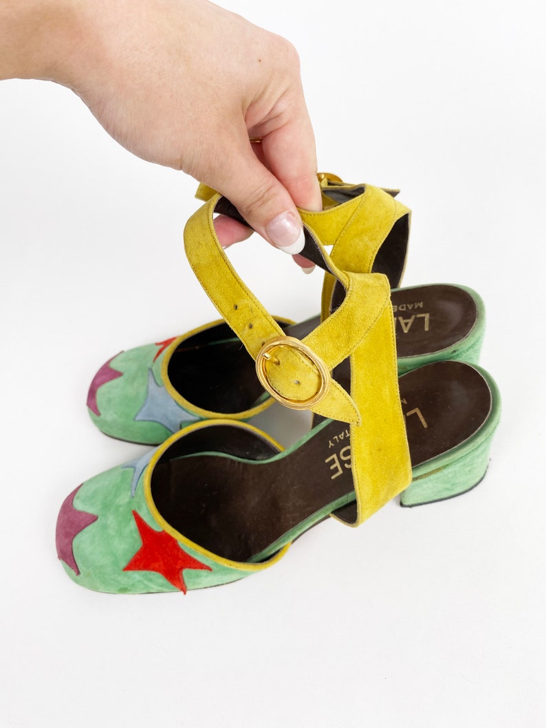 May include: Green suede heeled sandals with a yellow ankle strap and a star design. The sandals are made in Italy and have a brown leather insole. The brand name "L'AISE" is printed on the insole.