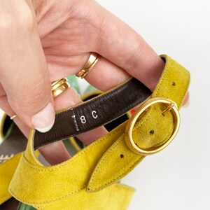 May include: Close-up of a yellow suede sandal with a gold buckle. The sandal has a brown leather strap with the size "8C" printed on it.