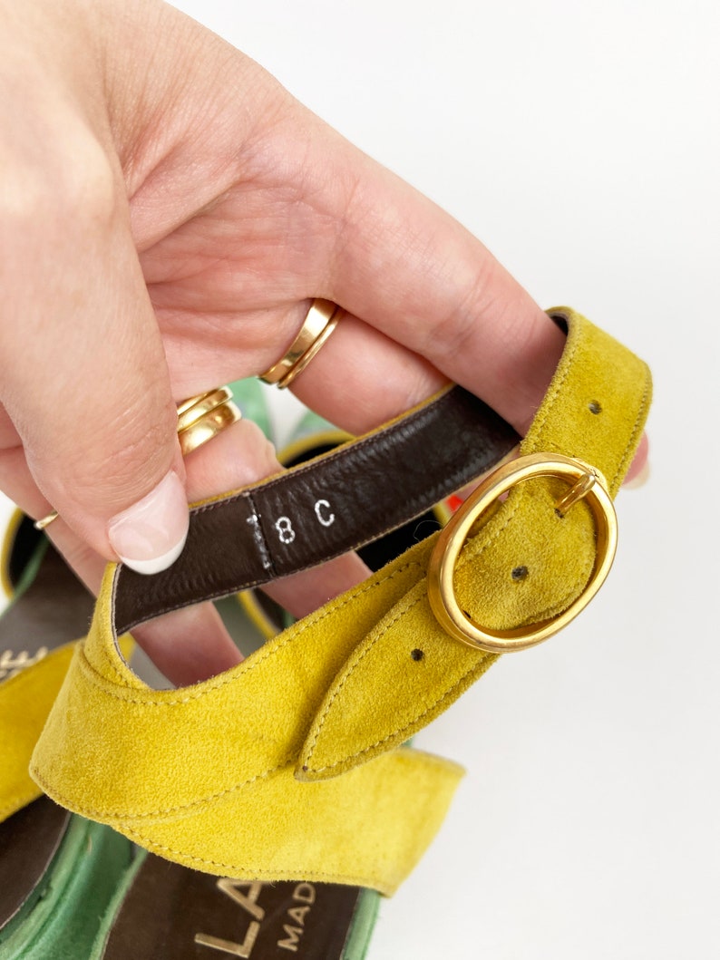 May include: Close-up of a yellow suede sandal with a gold buckle. The sandal has a brown leather strap with the size "8C" printed on it.