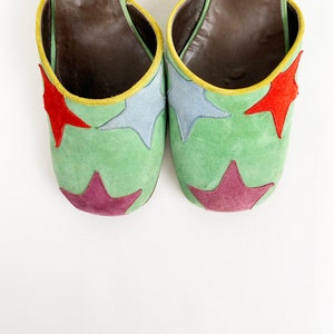 May include: A pair of green suede heeled shoes with colorful star accents. The shoes have a brown leather lining and a yellow trim around the edge.