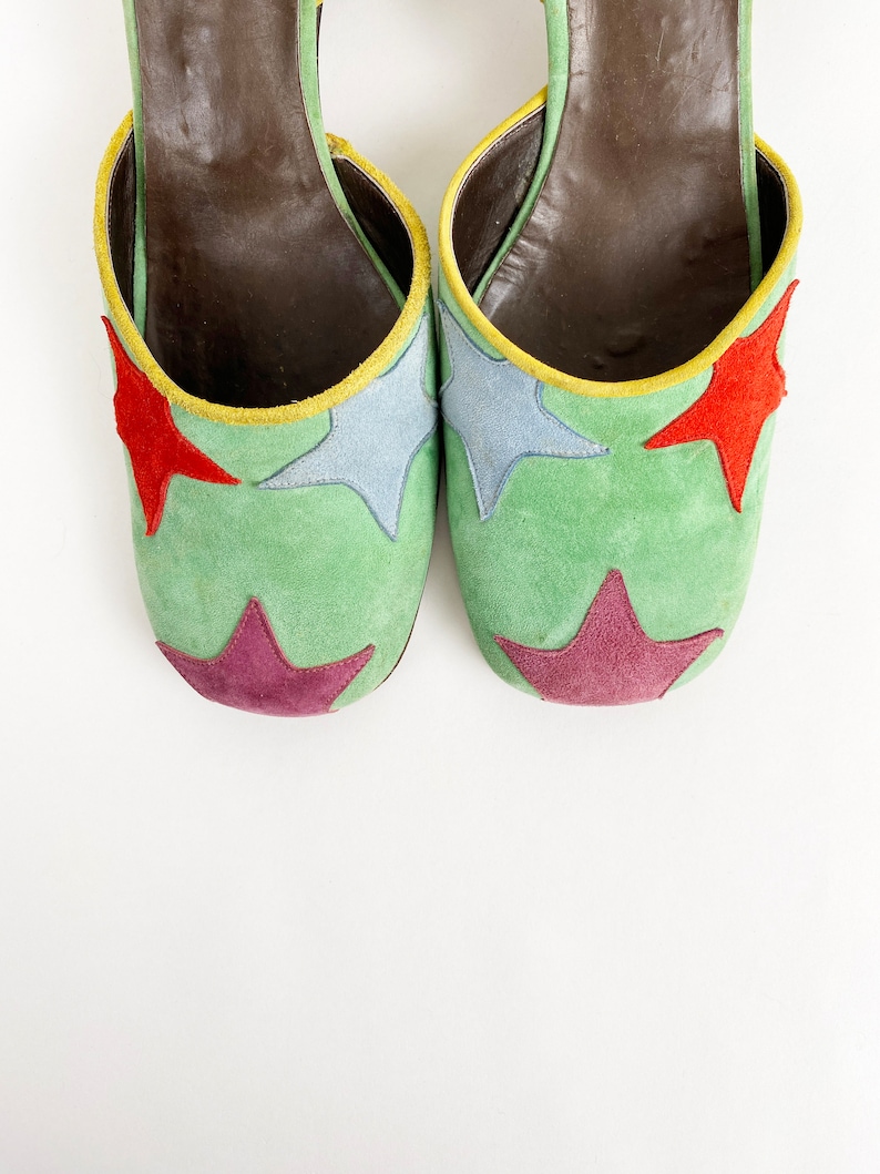 May include: A pair of green suede heeled shoes with colorful star accents. The shoes have a brown leather lining and a yellow trim around the edge.