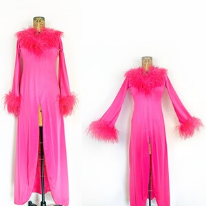 May include: A vibrant pink, floor-length gown with long sleeves and a high slit. The neckline and cuffs are adorned with fluffy pink feathers. The dress is displayed on a mannequin.