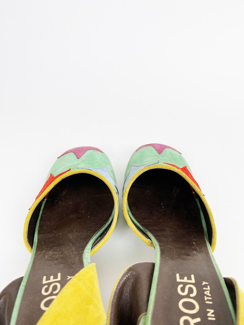 May include: A pair of yellow, green, blue, and red suede heeled sandals with a brown leather insole. The sandals have a star-shaped design on the top and the brand name "ROSE IN ITALY" is printed on the strap.