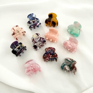 Puede incluir: Una colección de once pequeñas pinzas para el cabello en varios colores y patrones, incluyendo rosa translúcido, azul jaspeado, carey, melocotón sólido y verde menta. Cada pinza cuenta con un mecanismo de resorte metálico y dientes entrelazados.