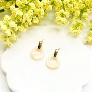 Könnte beinhalten: Ein Paar goldene Creolen mit einem weißen muschelförmigen Design. Die Ohrringe liegen auf einem weißen Hintergrund.