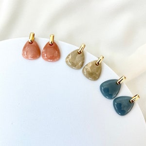 May include: A set of six earrings with gold posts and different coloured, marbled, teardrop-shaped stones. The stones are in shades of peach, brown, and blue.