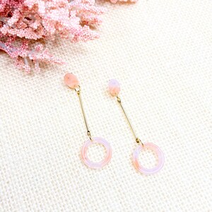 Könnte beinhalten: Ein Paar zarte Ohrringe mit einem pink-weißen Wirbelmuster. Die Ohrringe haben oben einen kleinen pink-weißen Kreis und unten einen größeren pink-weißen Kreis, die durch einen dünnen goldenen Draht verbunden sind.