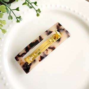 May include: Rectangular hair clip with a tortoiseshell pattern in shades of brown and cream. The clip features a gold-colored metal clasp. The hair accessory is designed to hold hair in place and add a touch of style.