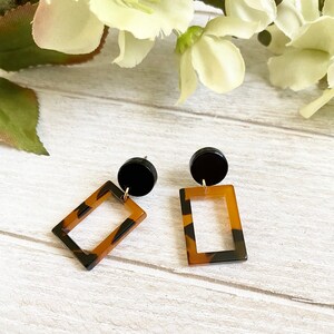 Könnte beinhalten: Ein Paar Ohrhänger mit einem einfarbigen schwarzen runden Stecker, der mit einem rechteckigen Rahmenanhänger mit braun-schwarzem Schildpattmuster verbunden ist. Die Ohrringe sind mit einer glatten, polierten Oberfläche und goldfarbenen Metallteilen gefertigt.