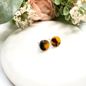 May include: A pair of small, round, brown and yellow tortoise shell stud earrings. The earrings are on a white surface.