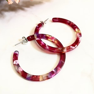 May include: A pair of large hoop earrings made of red, pink, and white marbled plastic. The earrings have a silver post and back.
