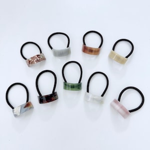 May include: A collection of eight hair ties with black elastic bands and rectangular, marbled-look accents in various colors. The accents include shades of brown, gray, green, and pink, offering a range of options for accessorizing hairstyles.