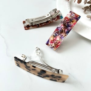 May include: Three hair clips with different patterns. One clip is brown with black spots, another is a rectangle with a floral pattern, and the last is a rectangle with a blue, pink, and white pattern.